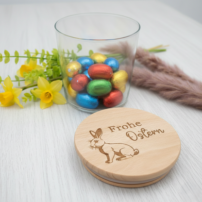 Ein hochwertiges Vorratsglas gefüllt mit bunten Ostereiern, davor lehnt ein runder Holzdeckel mit einer feinen Lasergravur eines Hasen und dem Schriftzug „Frohe Ostern“. Im Hintergrund sind rosa Tulpen auf einem dunklen Holztisch zu sehen – moderne Osterdekoration und Geschenkidee.